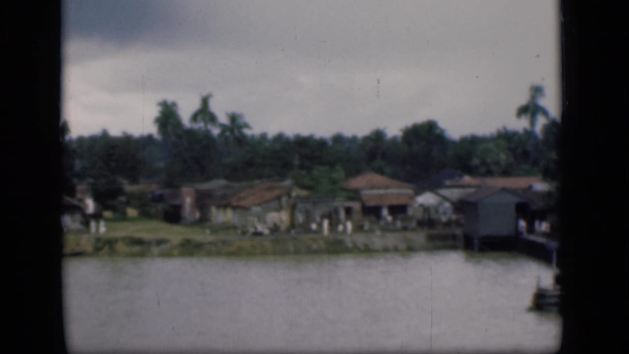 1946 Bombay 8mm frame showing low riverside houses, palms, and calm water under an overcast sky.