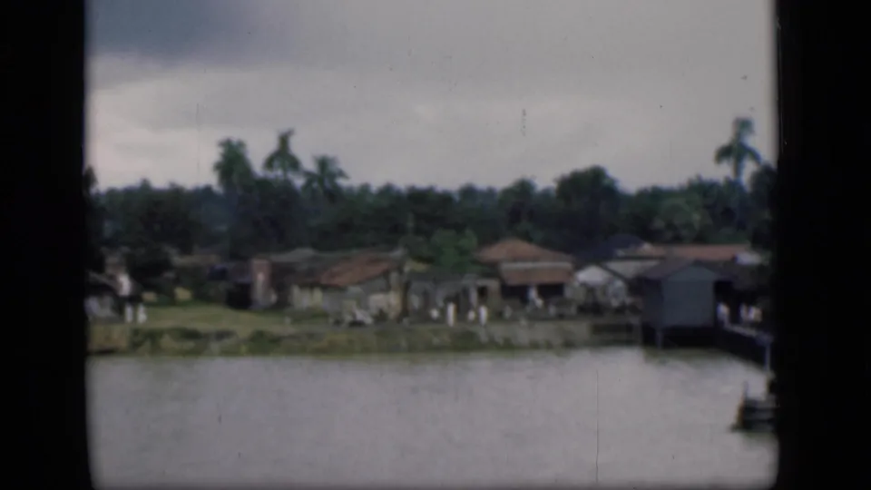 1946 Bombay 8mm frame showing low riverside houses, palms, and calm water under an overcast sky.