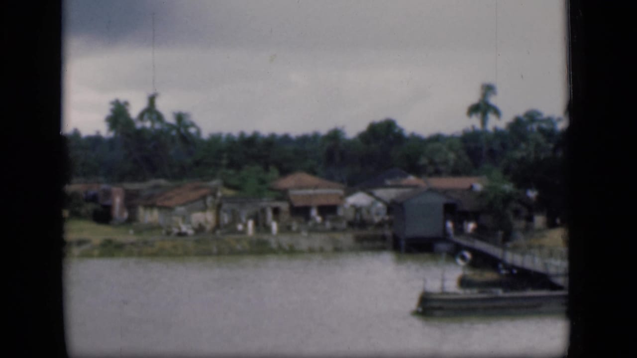 1946 Bombay riverside view with low homes, palms, a pier structure, and a moored boat near the frame edge.
