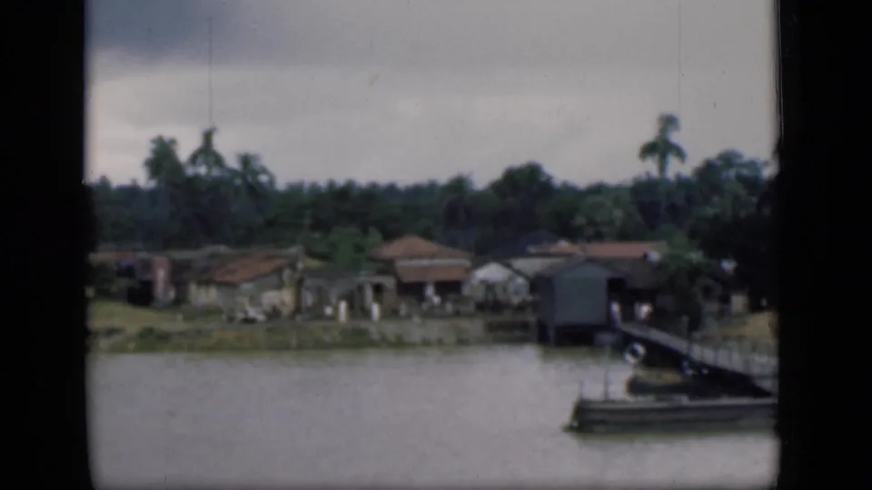1946 Bombay riverside view with low homes, palms, a pier structure, and a moored boat near the frame edge.