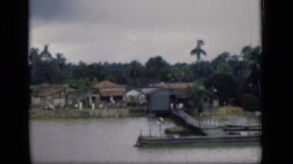 1946 Bombay home-movie frame with a wooden pier, moored boat, riverside houses, and palms across calm water.