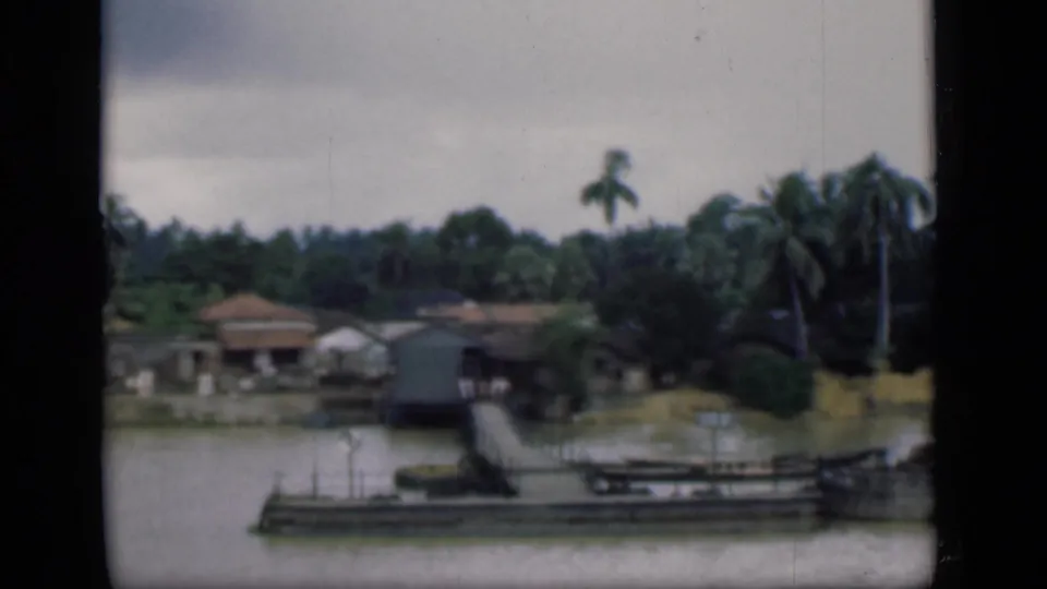 1946 Bombay riverbank frame showing a long moored boat, pier railings, low houses, palms, and dense greenery.
