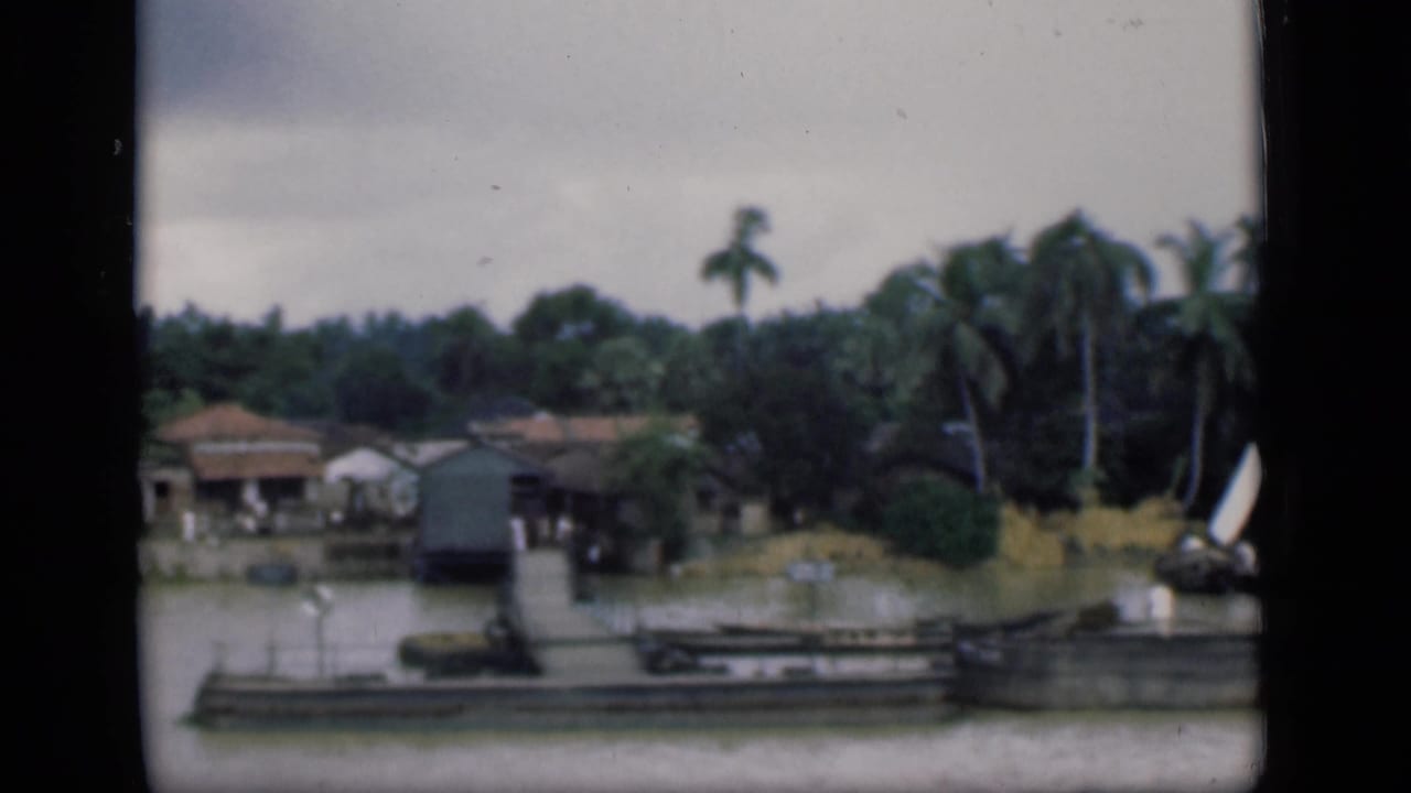 1946 Bombay home-movie frame showing low houses by water, a pier, coconut palms, greenery, and a small white-sailed boat.