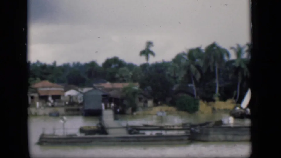 1946 Bombay home-movie frame showing low houses by water, a pier, coconut palms, greenery, and a small white-sailed boat.