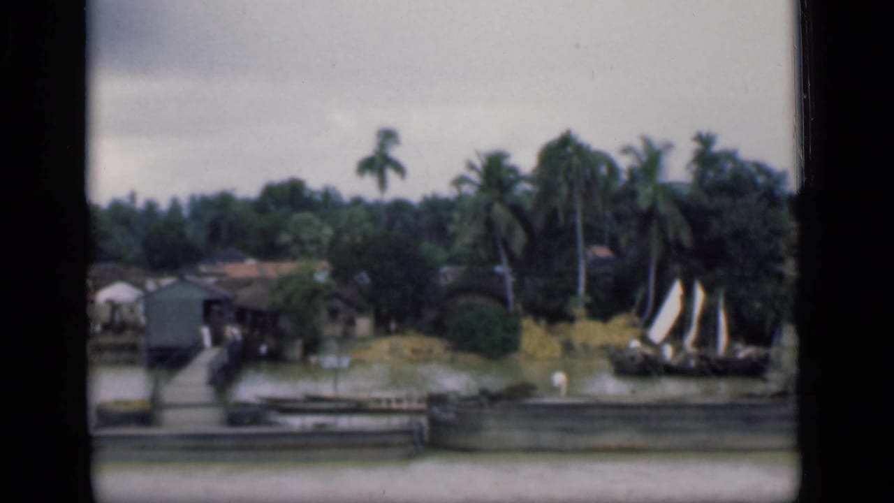 1946 Bombay footage frame with waterfront houses, wooden piers, coconut palms, calm water, and a small sailboat near the shore.