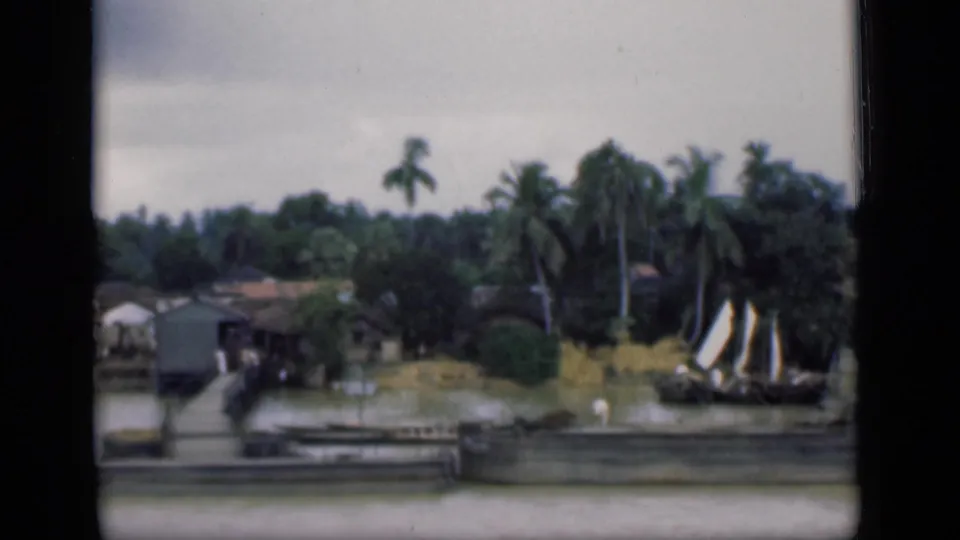 1946 Bombay footage frame with waterfront houses, wooden piers, coconut palms, calm water, and a small sailboat near the shore.