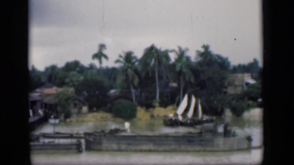 1946 Bombay home-movie frame showing a small sailboat passing near riverside homes, palm trees, piers, and dense greenery.
