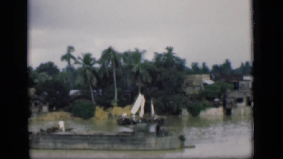 1946 Bombay frame with a small white-sailed boat on the water, backed by coconut palms, shoreline houses, greenery, and a low wall.