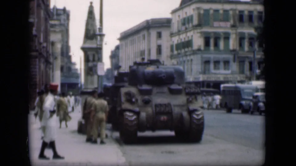 Bombay street scene from 1946 showing pedestrians, older cars, multi-story buildings, and a large armored-looking vehicle in the road.