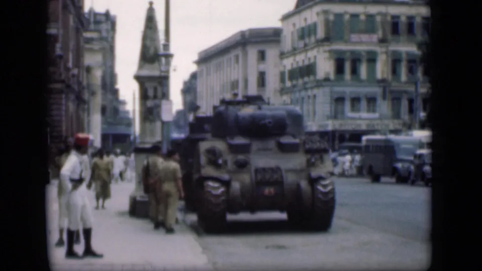 1946 Bombay street view with a tank at the curb, uniformed men on the sidewalk, pedestrians, older cars, and a tower-like monument in the background.