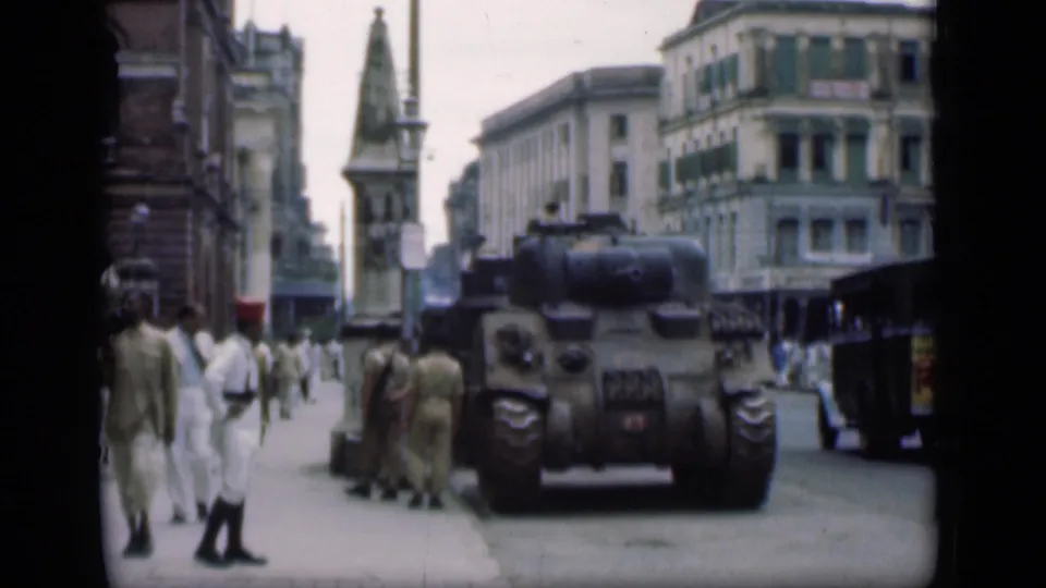1946 Bombay street scene showing a tank beside the curb, several uniformed men, pedestrians in white clothing, cars, and city buildings.