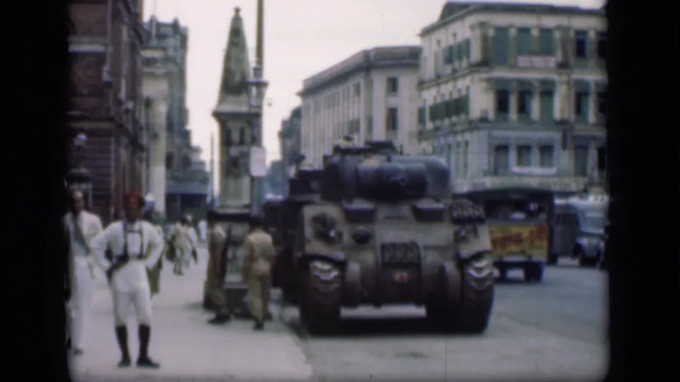 1946 Bombay footage with a tank parked near the street, a uniformed man in white in the foreground, soldiers by the curb, and passing vehicles.