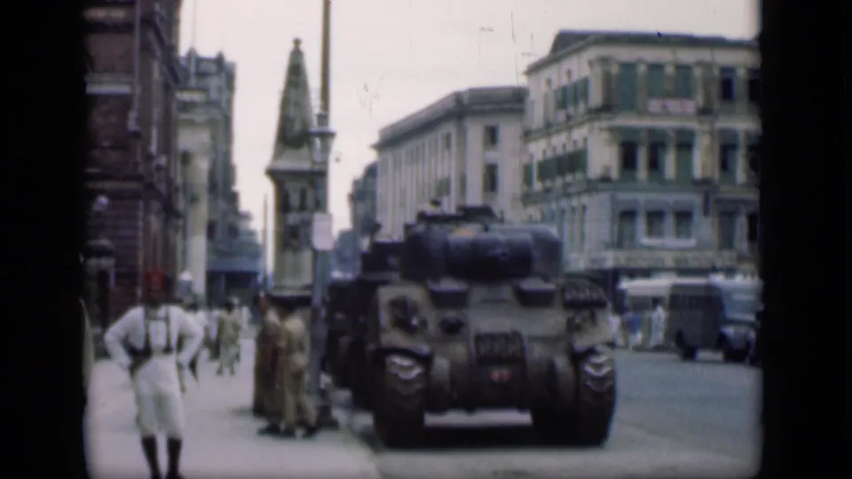 1946 Bombay city-center view with a tank, uniformed men on the sidewalk, pedestrians, older cars, and a tower or monument in the distance.