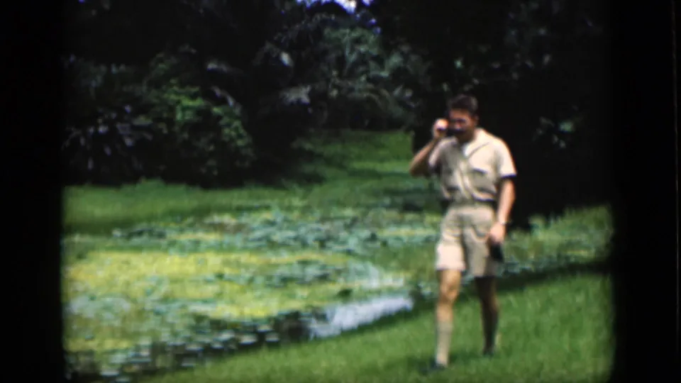 1946 Bombay frame showing an adult man in shorts and a light shirt walking along the grassy edge of a pond.