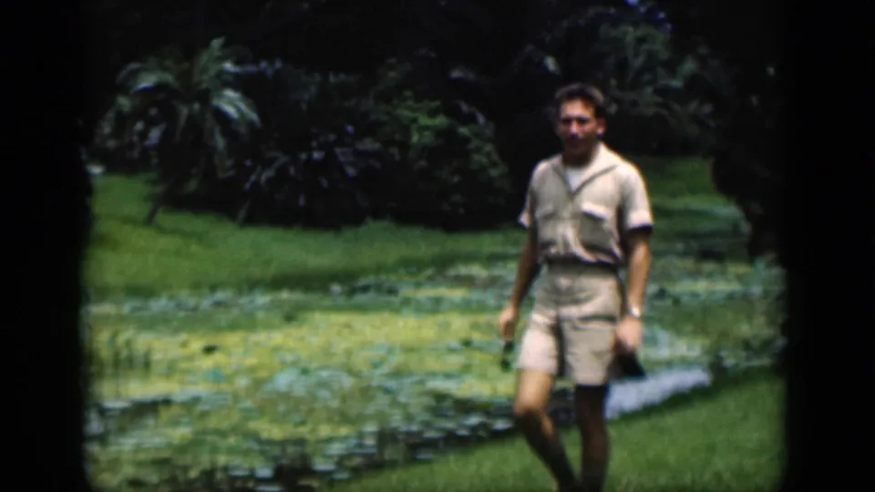 The man continues walking through the Bombay garden, with lily pads, grass, palms, and dark foliage visible behind him.