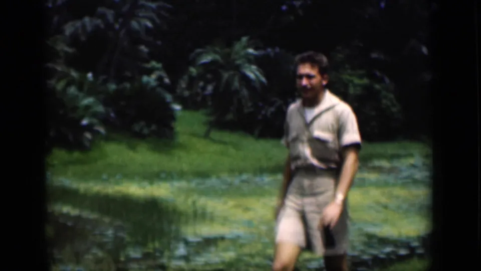 A closer 1946 Bombay home-movie frame shows the man walking near the pond, with palms and garden shrubs filling the background.