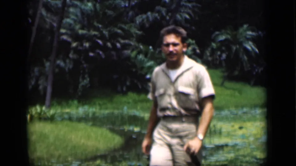 The man passes nearer the camera in the Bombay garden, holding a small dark object with the lily-pad pond and palms behind him.