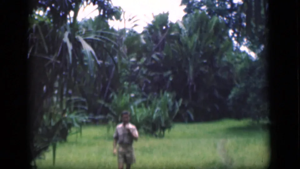 1946 Bombay home-movie frame with an adult man in light clothing emerging across grass beneath tall palm-like foliage.