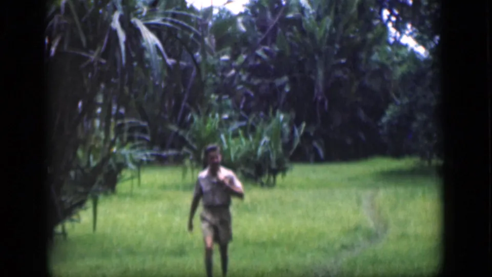 1946 Bombay frame showing an adult man in a light shirt and shorts walking toward the camera across a grassy clearing.