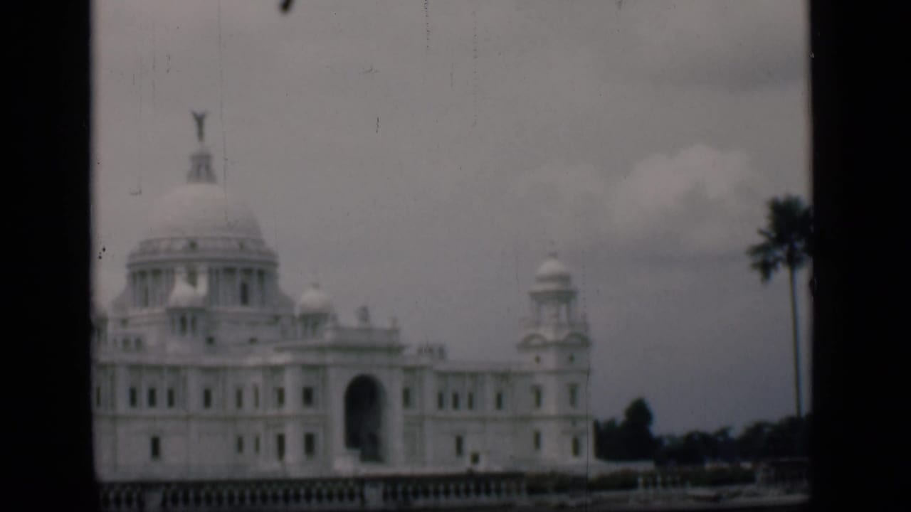 White domed public building with arched entrance, palm tree, cloudy sky, and dark film-frame edges.