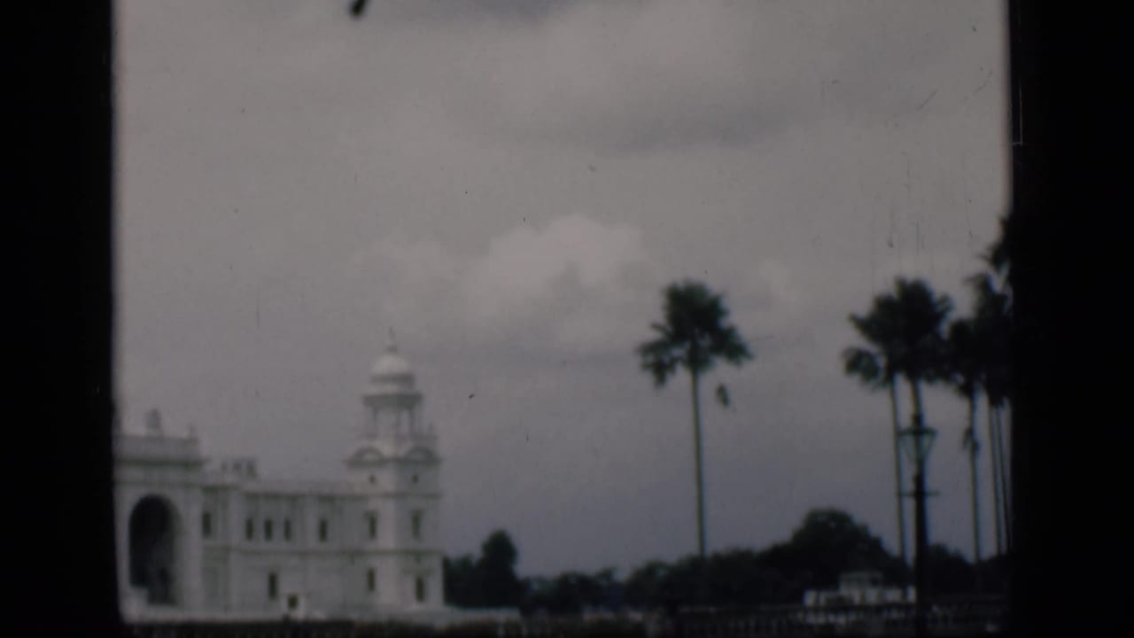 1946 8mm view identified as Bombay, India, showing a white landmark building partly framed by palms and dark window edges.