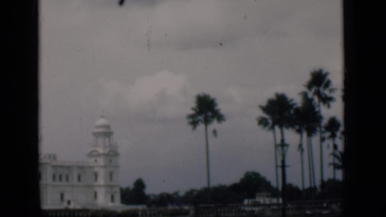 1946 8mm travel frame identified as Bombay, with a white towered landmark, palms, and cloudy sky seen from a framed viewpoint.