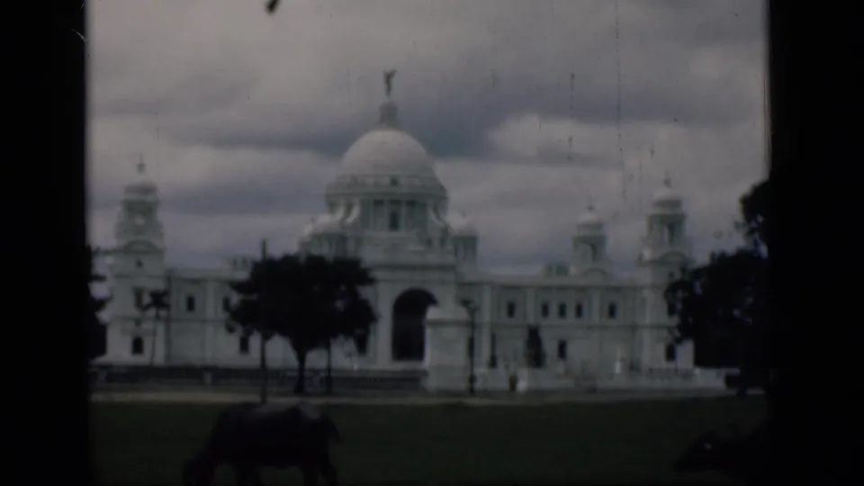 1946 8mm frame identified as Bombay, India, showing a domed white monument across a lawn with trees and a cow in front.