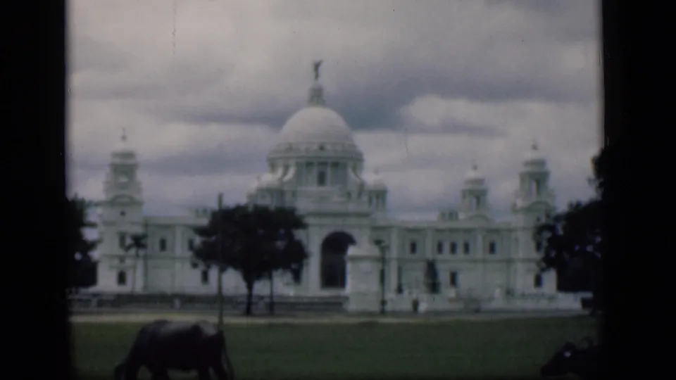 8mm frame of the Victoria Memorial exterior under clouds, with dark trees and buffalo grazing along the lawn.