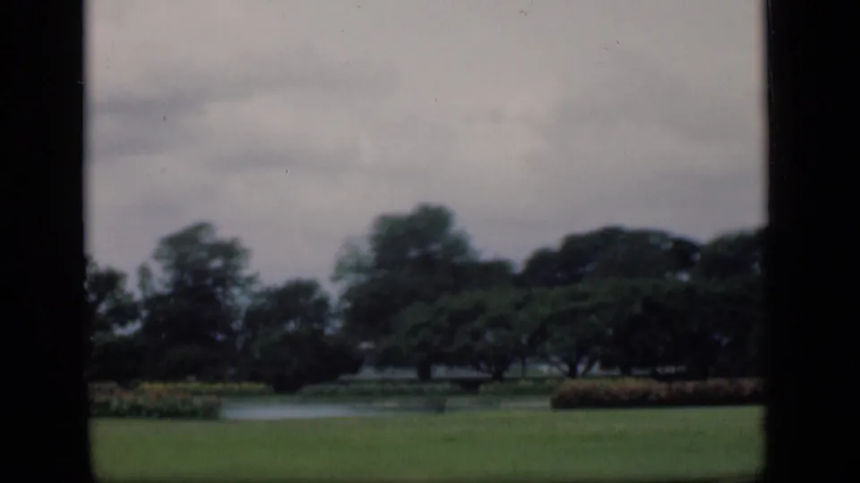 1946 Bombay home-movie frame of a landscaped park, showing lawn, pond, hedges, and a dense tree line.
