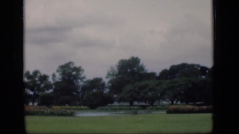 1946 8mm view of a Bombay garden with lawn, water, flowerbeds, and mature trees under an overcast sky.