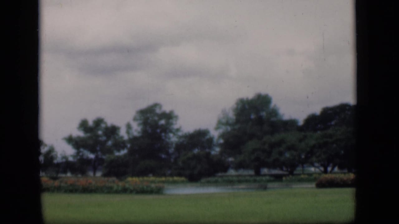 Bombay garden scene on 8mm film, showing a clipped lawn, pond, flowerbeds, and trees with muted color.