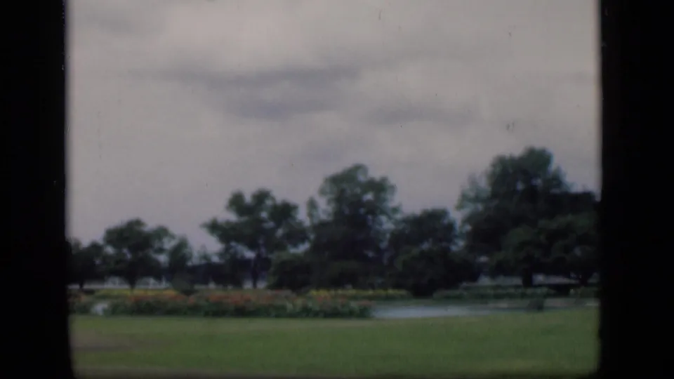 Final 8mm frame of the Bombay garden view, with lawn, pond, flowerbeds, trees, and gray sky framed by dark borders.
