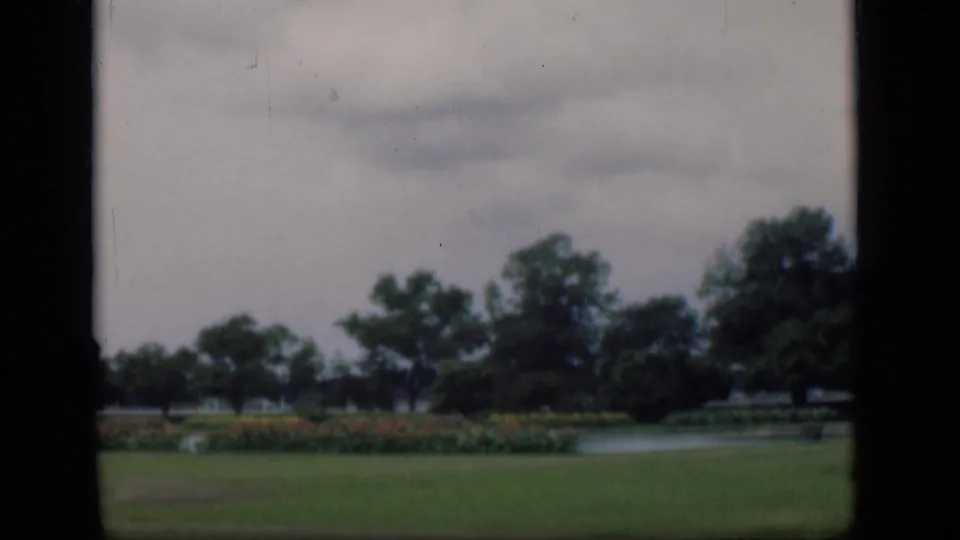 1946 Bombay park view with lawn, flowering bushes, mature trees, and a small lake under an overcast sky.