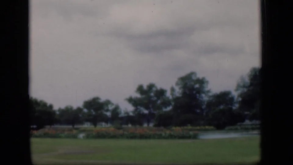 The pan continues across a Bombay garden scene, showing open grass, low flowers, water, and tree silhouettes.