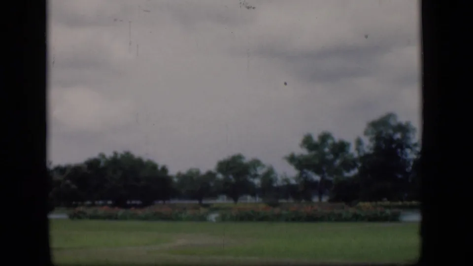 A later frame in the Bombay park pan shows lawn, flowering beds, mature trees, and a muted lake view beneath cloudy skies.