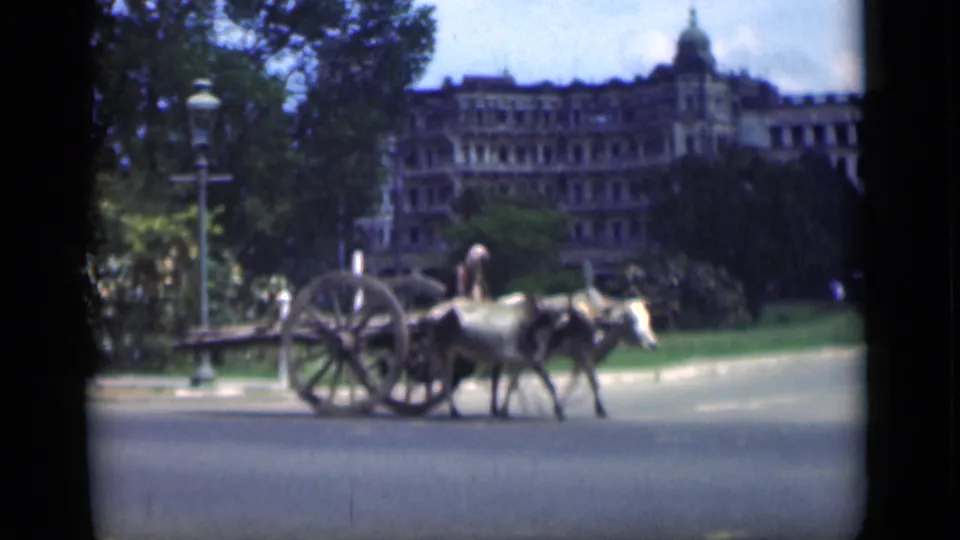 1946 Bombay home-movie frame of an ox-drawn cart moving past lawns, trees, and a large colonial-style building.