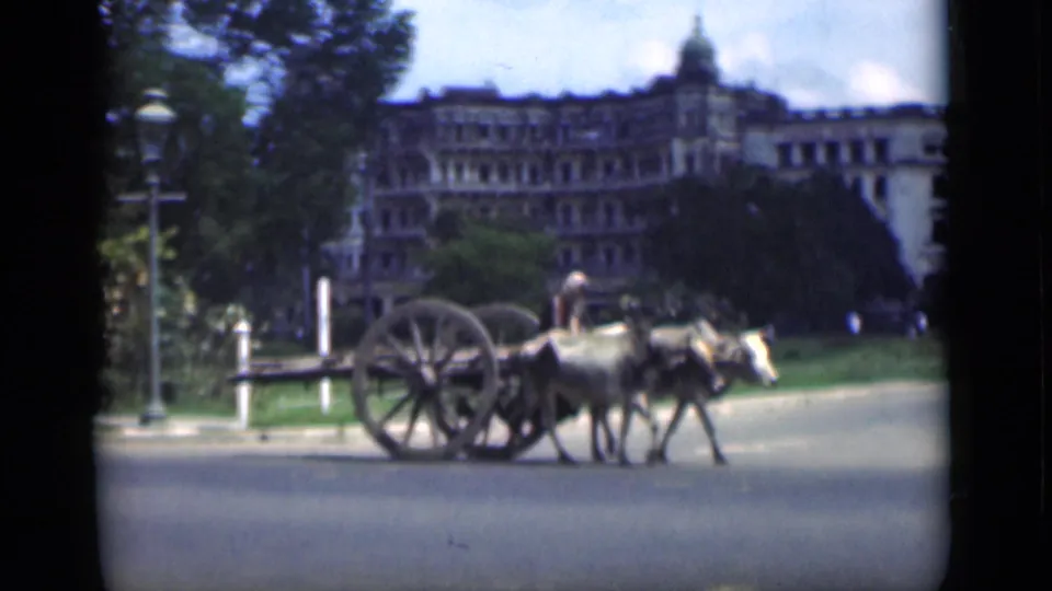1946 Bombay home-movie frame showing a handler guiding an animal-drawn cart past trees, lampposts, and a large colonial-era building.