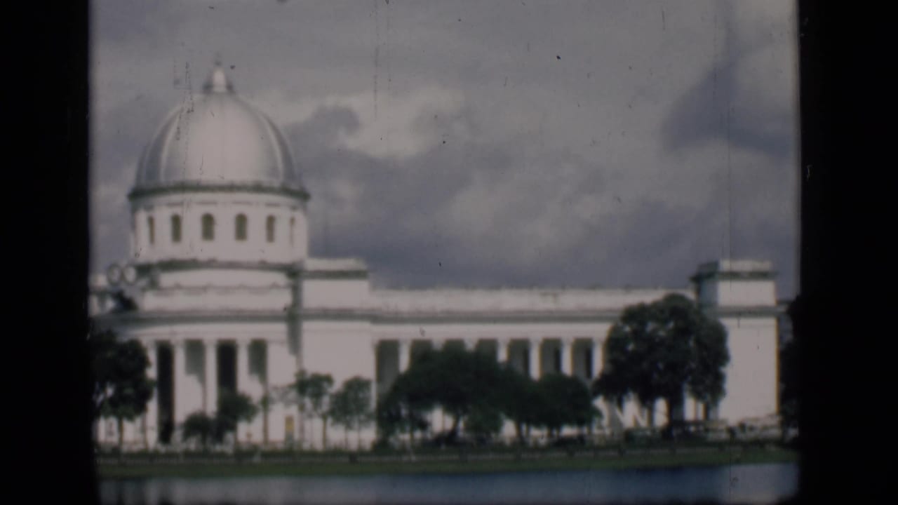 1946 Bombay home-movie view of a white domed civic building across water, partly framed by trees under a cloudy sky.