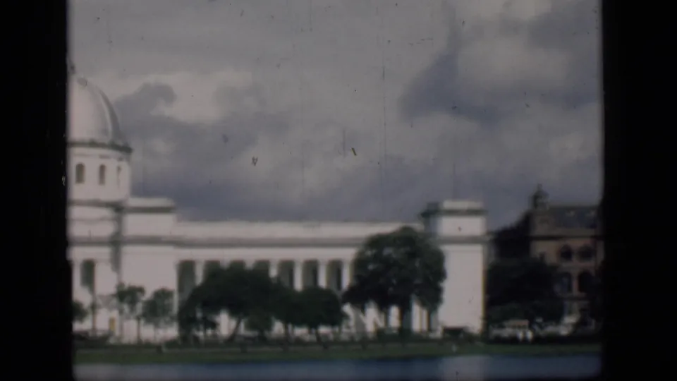 Soft 8mm frame from Bombay showing a partial white civic building, waterfront foreground, trees, and small cars at the roadside.