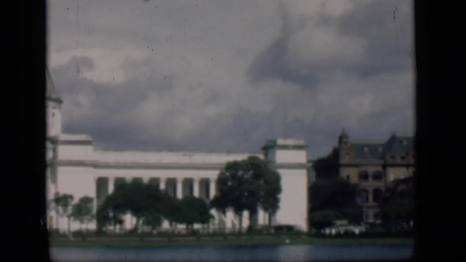 1946 Bombay view of a white civic facade across water with trees, adjacent buildings, and small automobiles beneath a cloudy sky.
