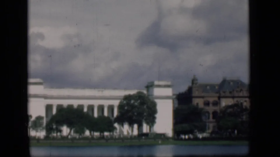 1946 Bombay view across water toward a white columned building, trees, and a darker multi-story structure under clouds.