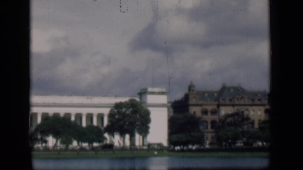 Waterfront buildings with trees, a white facade, and cloudy sky within dark film borders.