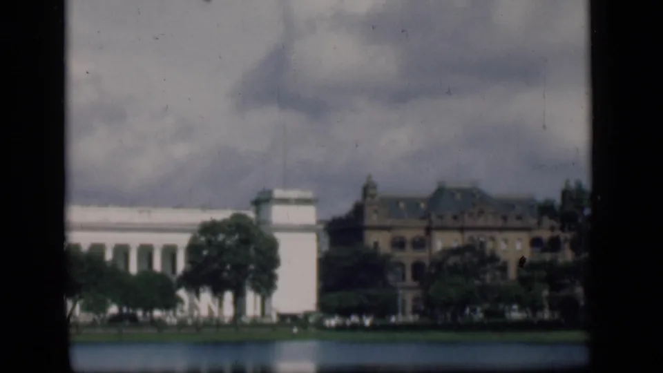 Soft 1946 Bombay 8mm frame showing a white columned waterfront building, trees, water, and a brown structure nearby.