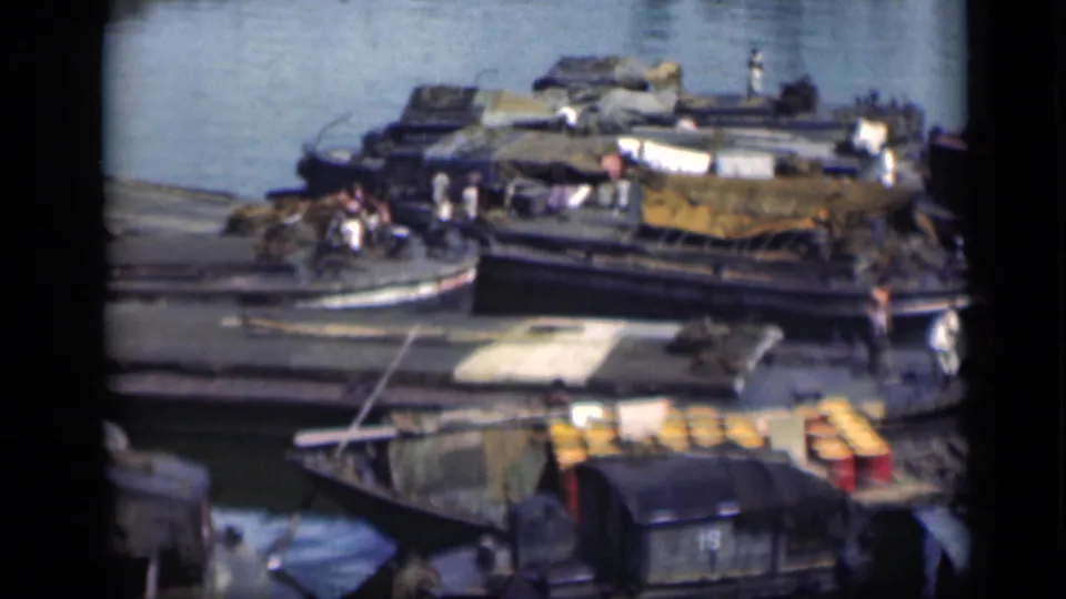 1946 Bombay port view with stacked yellow barrels, covered boat cargo, several wooden hulls, and small figures moving near the water.