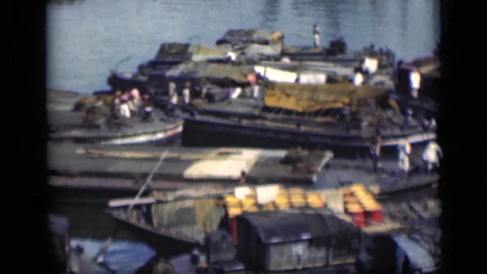 1946 Bombay waterfront footage showing crowded wooden boats, people aboard, tarped cargo, yellow barrels, and a grey shoreline structure.