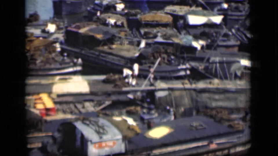 Bombay waterfront frame with distant figures, wooden boats, covered cargo, ropes or rails, and dark water around the docked vessels.