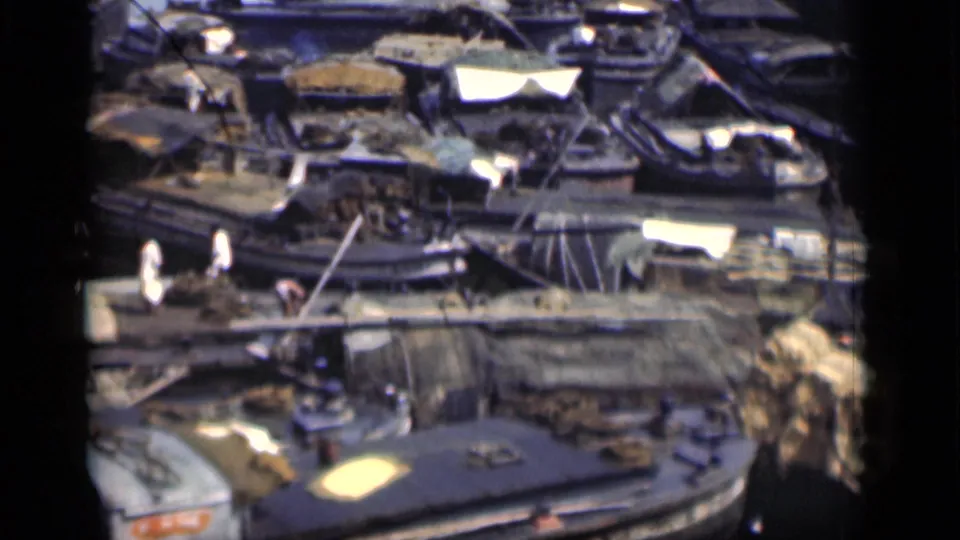 Crowded 1946 Bombay waterfront with pedestrians on a plank route, packed small boats, gear, and a blurred figure at the edge.