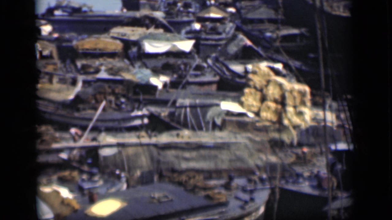 Crowded waterfront area with boats, tarps, stacked goods, and crane lines.