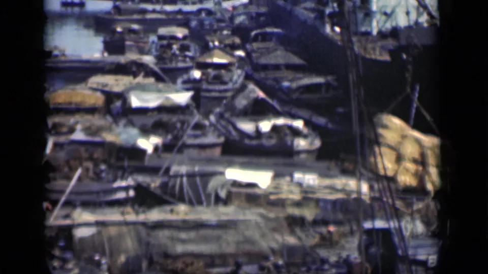 1946 Bombay home-movie frame showing a busy waterside settlement or work area with boats, tarps, stacked materials, and lines crossing the frame.