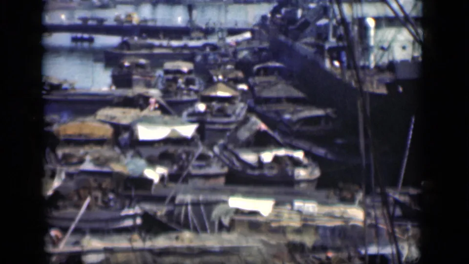 1946 Bombay frame showing small boats and covered goods packed among low waterside structures, with rigging or crane lines at the edge.